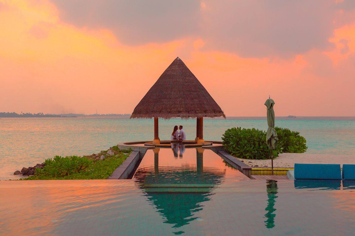Pareja bajo un quincho con techo de paja frente al mar al atardecer, en un resort